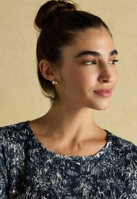 Top s námorným vzorom s strihanou textúrou, okrúhlym výstrihom a gombíkovými detailmi. Modelka má vlasy v bunke a nosí zlaté náušnice.