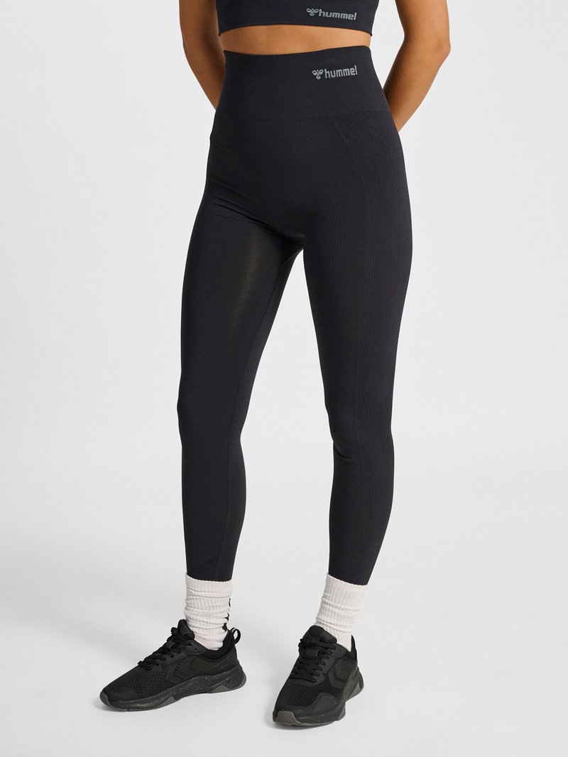 Vrouw die zwarte leggings met hoge taille draagt, zwarte sport-bh, witte sokken en zwarte sportschoenen, staand tegen een effen achtergrond.