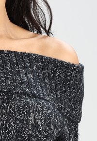 Suéter negro y gris de punto con caída de hombros que presenta un diseño de canalé grueso y hilos más claros en contraste para aportar textura.