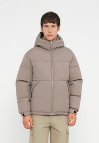 Jachetă tip puffer dintr-un material gri texturat, având un fermoar frontal, glugă și un design matlasat. Accente întunecate la mansete, asortate cu pantaloni cargo bej.