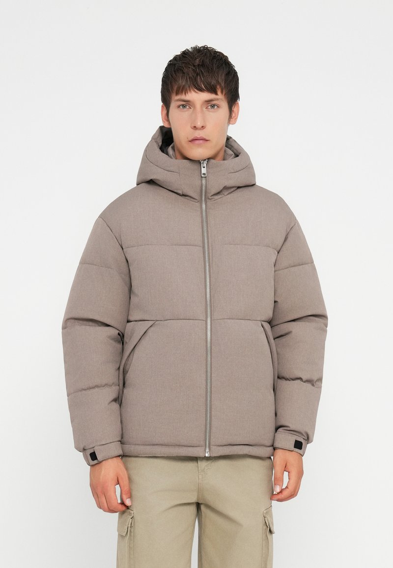 Jachetă tip puffer dintr-un material gri texturat, având un fermoar frontal, glugă și un design matlasat. Accente întunecate la mansete, asortate cu pantaloni cargo bej.
