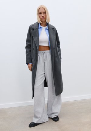 Un long manteau gris par-dessus un crop top blanc, associé à un pantalon de survêtement gris et des mocassins noirs, avec une chemise bleu clair en dessous.