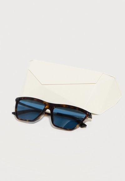 Ορθογώνια γυαλιά ηλίου με ταρταρούγα και μπλε φακούς τοποθετημένα μπροστά από μια γωνιώδη λευκή θήκη γυαλιών σε λευκή επιφάνεια.