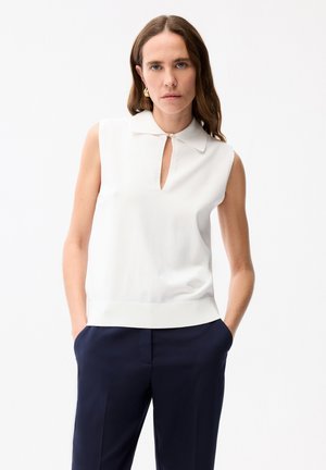 Femme portant un haut sans manches blanc à col avec une fente sur le devant et un pantalon bleu marine, debout les mains dans les poches devant un fond blanc.