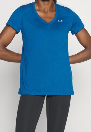 Persona que lleva una camiseta deportiva azul con cuello en V y un pequeño logo en el pecho, y leggings ajustados negros, brazos doblados con las manos en las caderas.