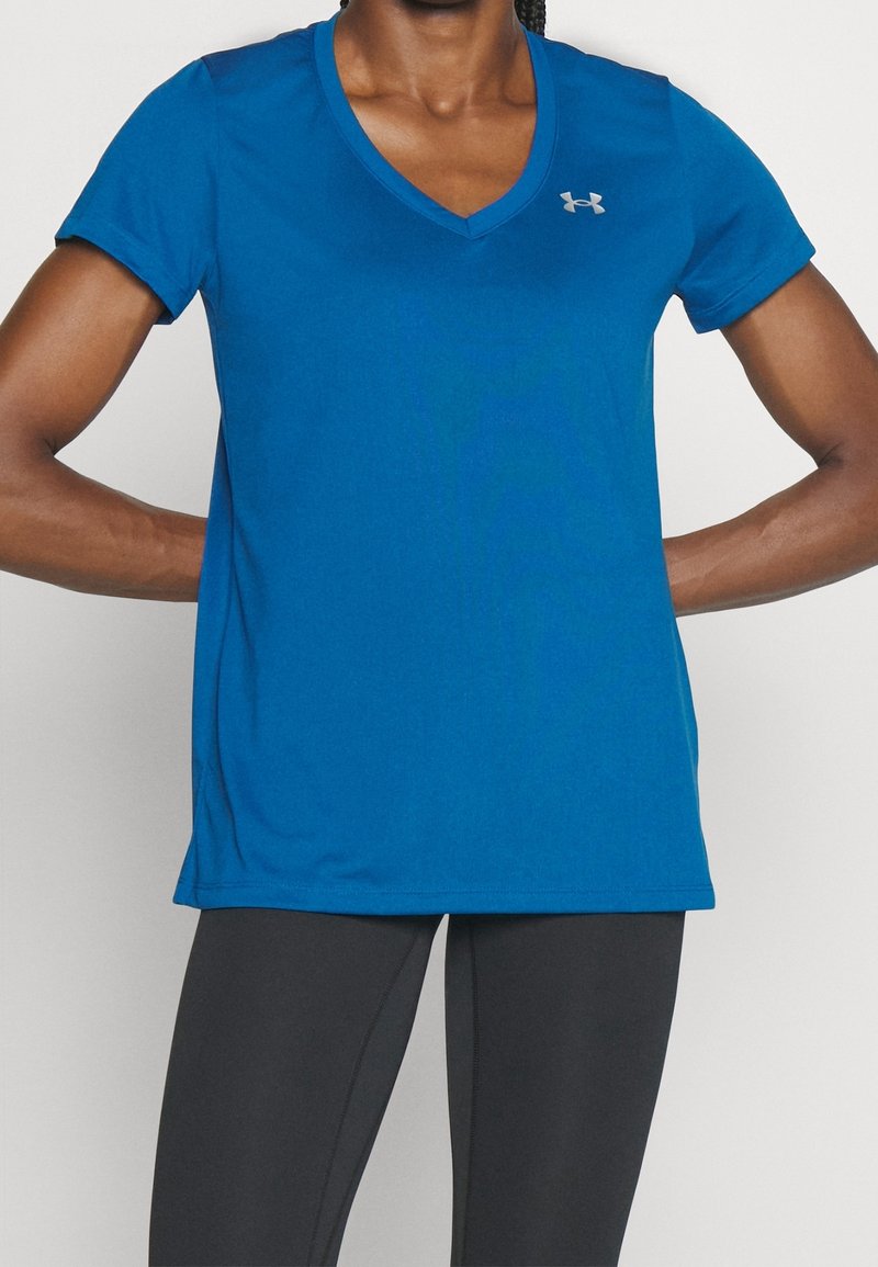 Personne portant un t-shirt de sport bleu à col en V avec un petit logo sur la poitrine, et des leggings noirs moulants, bras pliés avec mains sur les hanches.