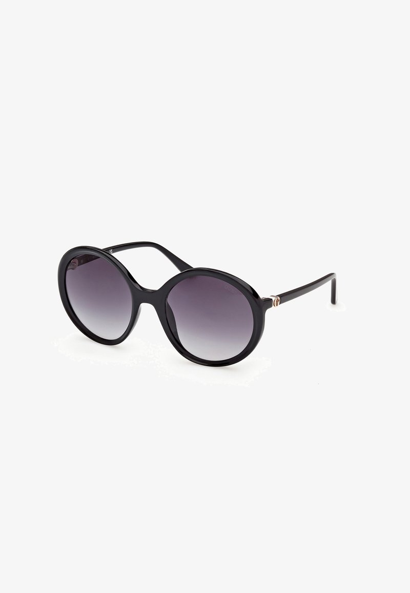 Lunettes de soleil noires oversize à monture ronde avec des verres dégradés foncés et des branches fines reposant sur une surface blanche.