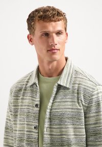 Gestreepte, button-up shirt in groene en witte stof met een textuurpatroon; gedragen over een effen groene crewneck t-shirt.