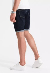 Pantalones cortos de mezclilla oscura con puños enrollados, que presentan dos bolsillos traseros y costuras amarillas, combinados con zapatillas blancas.