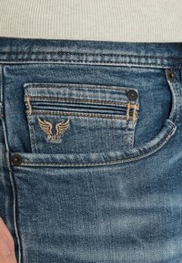 Jeans de denim en un tono azul desgastado, con un bolsillo trasero que presenta costuras verticales y un logotipo de alas bordado. Acentos de botones metálicos visibles.