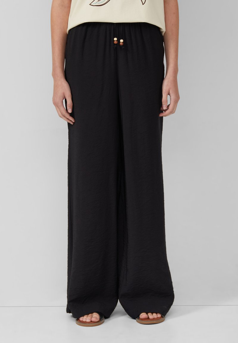 Pantalon noir à jambes larges en tissu léger, doté d'une ceinture élastique avec des perles décoratives, et d'une finition lisse et texturée.