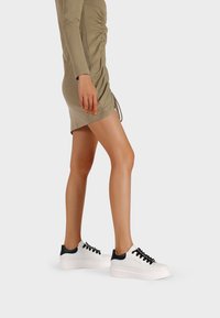 Sneakers bianchi con accentuazioni nere sul tallone e sui lacci. Presentano una superficie morbida e una suola spessa. Indossati con un vestito aderente color oliva.