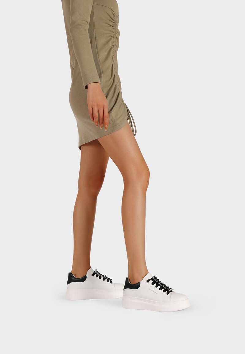 Sneakers bianchi con accentuazioni nere sul tallone e sui lacci. Presentano una superficie morbida e una suola spessa. Indossati con un vestito aderente color oliva.