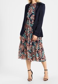 Blazer navy sopra un vestito floreale con motivi di foglie rosa e turchese, di lunghezza media, con vita arricciata e tacchi neri con cinturini.