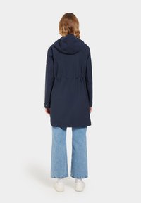 Marineblauer wasserdichter Mantel mit tailliertem Schnitt, Kapuze und langen Ärmeln. Kombiniert mit hellblauen weiten Jeans und weißen Turnschuhen.