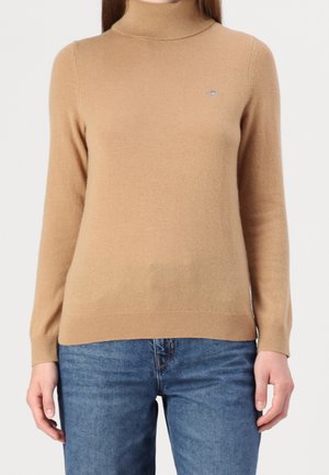 Kobieta ubraną w golf w kolorze wielbłądzim w połączeniu z niebieskimi dżinsami, stojąca na jednolitym tle.