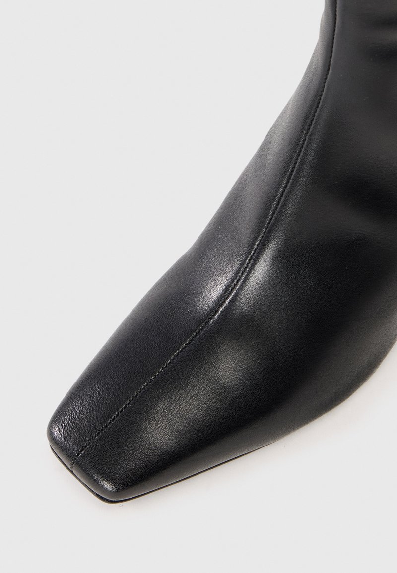 Czarny skórzany but z kwadratowym czubkiem i centralnym przeszyciem na jasnoszarym tle.