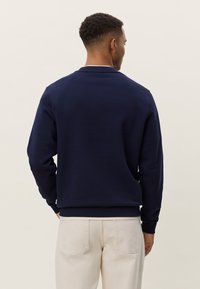 Temno modra jopa iz mehke tkanine. Ima okrogel izrez, rebraste manšete in spodnji rob ter nima vidnih vzorcev ali poudarkov.