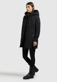 Kobieta w długim czarnym pikowanym puchowym płaszczu z kapturem, czarnych legginsach i czarnych masywnych butach, stojąca bokiem na białym tle.