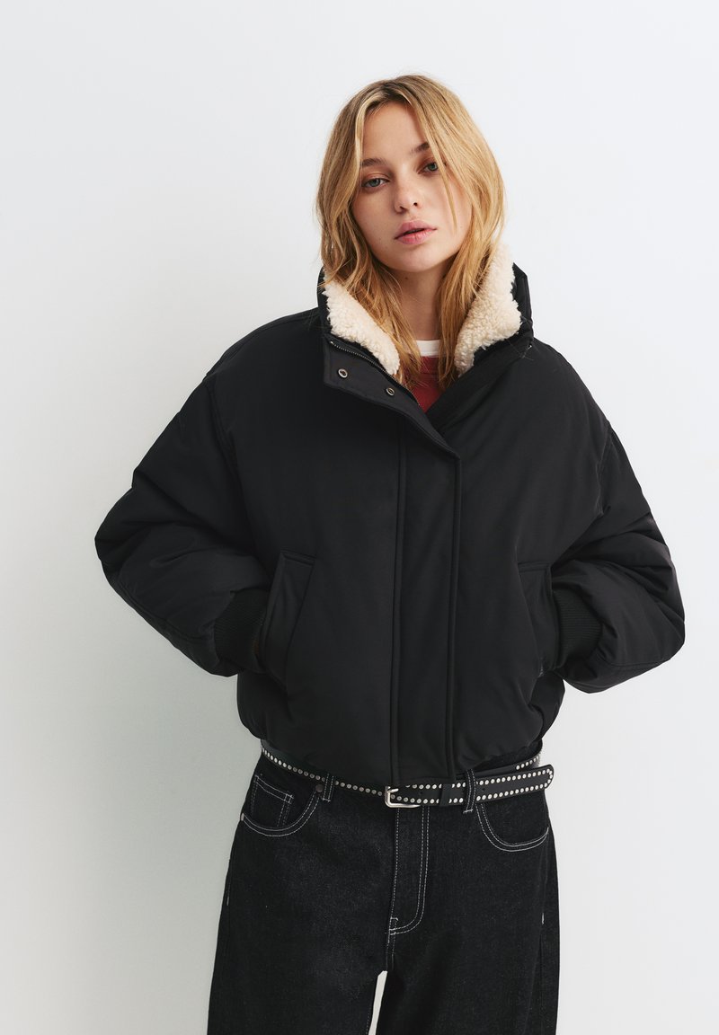 Černá péřová bunda s krémovým límcem z umělého shearlingu, má přední zip, boční kapsy a volný střih, nošena přes černé džíny.