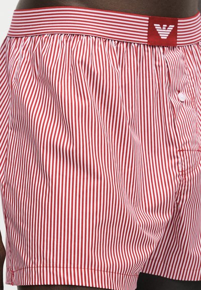 Άνδρας που φορά κόκκινα και λευκά κάθετα ριγωτά μποξεράκια με ελαστική μέση με κόκκινο ταμπελάκι και λευκό λογότυπο.