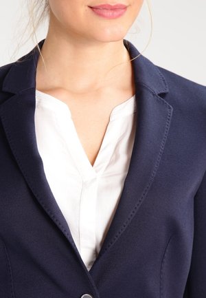 Kvinde iført en hvid bluse og en mørkeblå blazer, der viser kraven og det øverste af brystområdet.