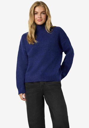 Kungligt blå tröja med hög ribbad hals, med en texturerad övre del och slät tyg nedanför. Matchas med svarta jeans.