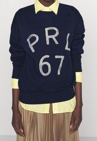 Navy sweatshirt with white cracked print "PRL 67," paired with a light yellow collared shirt, over a pleated beige skirt.