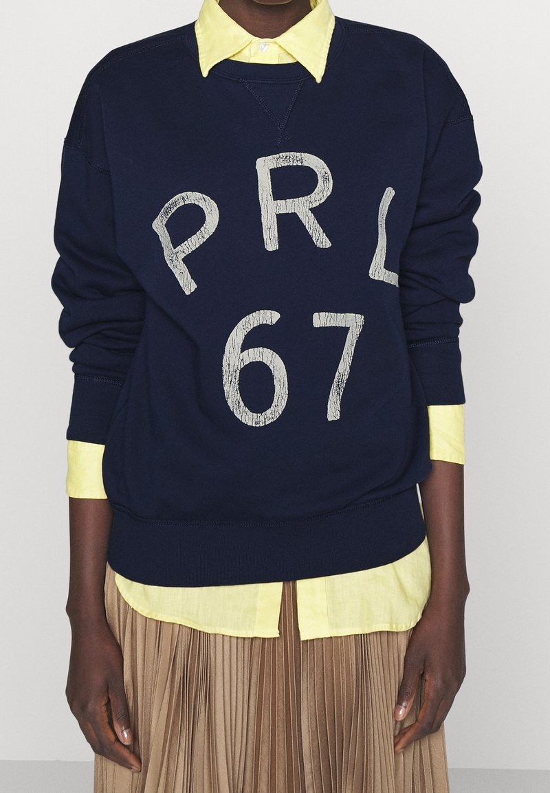Navy sweatshirt with white cracked print "PRL 67," paired with a light yellow collared shirt, over a pleated beige skirt.