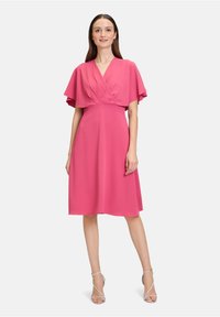 Rosa Kleid mit Wickeldesign und fließenden Ärmeln, knielang, aus leichtem Stoff. Kombiniert mit hohen Schuhen mit transparenten Riemen.