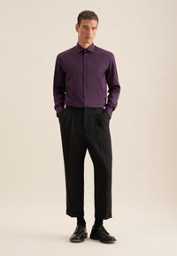 Camicia viola a maniche lunghe con chiusura a bottoni, indossata con pantaloni neri a pieghe e scarpe di pelle nere, in piedi su uno sfondo neutro.