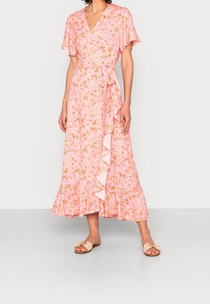 Femme portant une robe portefeuille rose à motif floral avec un ourlet à volants et des manches courtes évasées, assortie à des sandales beiges à enfiler.