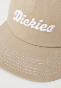 Barna baseball sapka fehér hímzett "Dickies" logóval, texturált anyagból, ellentétes fehér varrással a karimán.