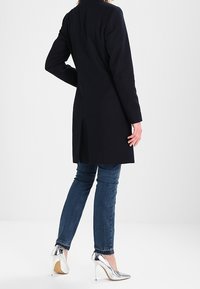 Czarny wełniany płaszcz z długimi rękawami, prostym krojem i rozporkiem z tyłu, zestawiony z niebieskimi dżinsami i srebrnymi szpilkami.