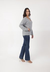 Szary sweter robiony na drutach z dekoltem w kształcie litery V, w zestawieniu z ciemnoniebieskimi dżinsami w dzwony i przezroczystymi sandałami na obcasie. Miękka tekstura, luźny krój.