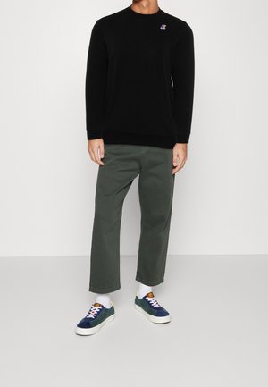 Man met zwarte longsleeve met klein logo, groene cropped broek, witte sokken en blauwe sneakers op een grijze vloer met een witte muur als achtergrond.