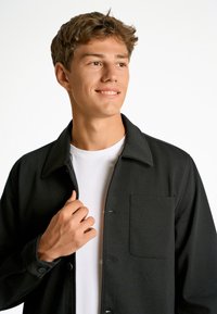 Chaqueta negra de botones con cuello, que cuenta con un bolsillo frontal y una textura suave, llevada sobre una camiseta blanca.