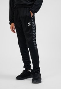 Czarne sportowe spodnie o delikatnym połysku, z elastycznym pasem, bocznymi kieszeniami oraz wzorem w jodełkę w kolorze szarym na nogawkach.