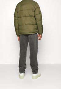 Oliwkowa, puchowa kurtka z pikowanym wzorem, noszona na szarych spodniach dżinsowych. Charakteryzuje się czystym wykończeniem i ściągaczami na mankietach, uzupełniona butami z jasną podeszwą.