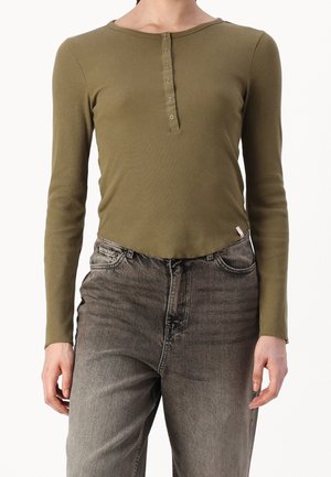Osoba ubrana w oliwkowo-zieloną, prążkowaną koszulę z długim rękawem i zapinanym na guziki klinem oraz wyblakłe czarne, wysokich stanem dżinsy, stojąca na białym tle.