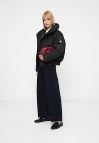Doudoune noire avec un col montant et une fermeture éclair sur le côté, associée à un jean noir à jambes larges et une pochette bordeaux. Texture lisse et branding minimal.