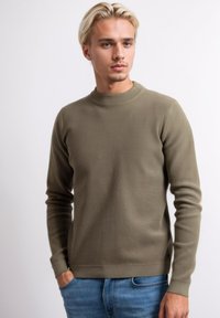 Khaki geribbelde trui met een ronde halslijn en lange mouwen. De stof lijkt textuur te hebben met een aansluitende pasvorm. Gecombineerd met blauwe jeans.