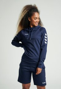 Mörkblå atletisk hoodie med snörning, långa ärmar och vita chevron-accenter. Matchas med mörkblå shorts. Mjuk tygstruktur.