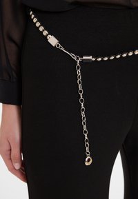 Zilveren riem met ronde studs, voorzien van een sluiting en kettingdetail dat naar beneden uitsteekt. Tegen een zwarte stof achtergrond.