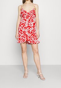 Rotes und weißes Blumen-Minikleid mit Rüschen, V-Ausschnitt und dünnen Trägern. Kombiniert mit weißen, schmalen High Heels vervollständigt es den Look.