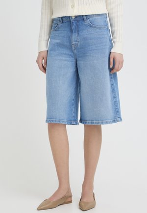 Kobieta nosząca jasnoniebieskie dżinsowe szorty do kolan, białą bluzkę z długim rękawem i beżowe spiczaste baleriny.