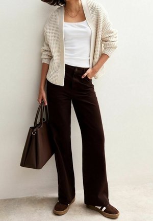 Kobieta ubrana w kremowy sweter na guziki, biały top na ramiączkach, ciemne szerokie spodnie, trzyma brązową torbę na ramię i ma na stopach brązowe wsuwane buty.