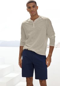 Maglietta henley a maniche lunghe color beige chiaro con bottoni abbinata a pantaloni corti blu scuri. Tessuto morbido; vestibilità comoda; senza motivi né grafiche.