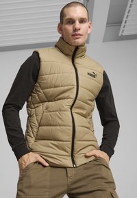 Gilet trapuntato beige con colletto alto, zip nera e logo Puma; abbinato a una maglietta nera a maniche lunghe e pantaloni kaki.
