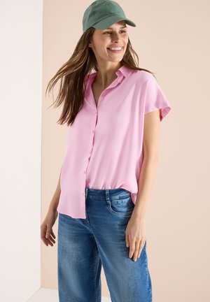 Smilende kvinde iført en grøn kasket, lyserød kortærmet skjorte med knapper og blå jeans mod en ensfarvet beige baggrund.